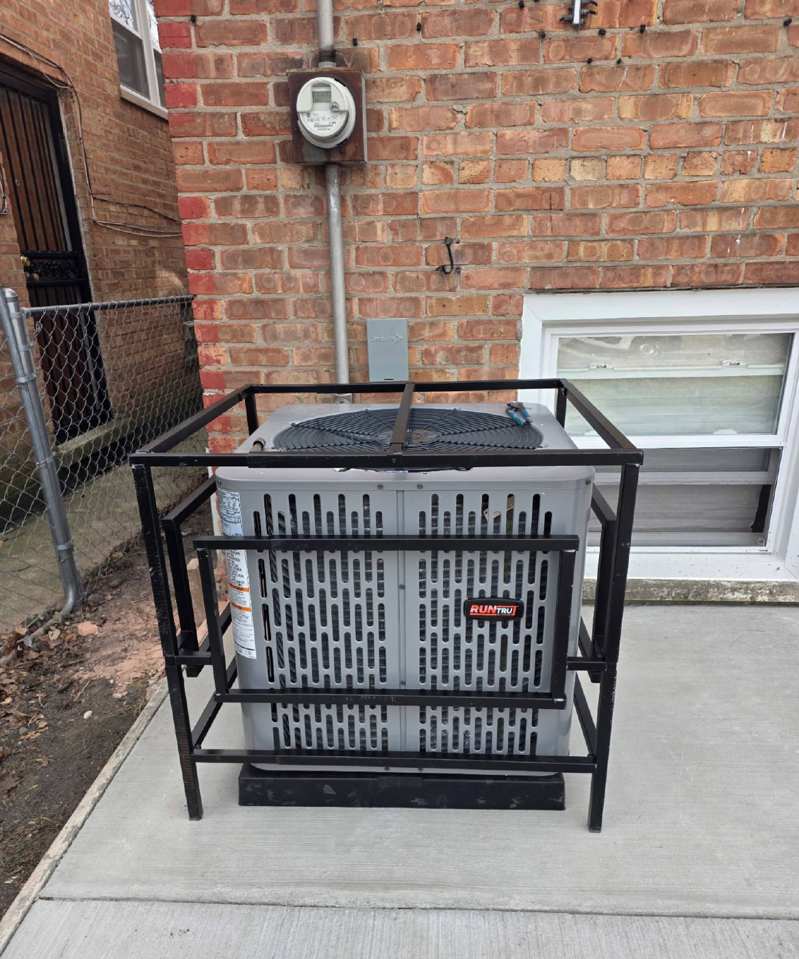 A new outdoor air conditioner on a fresh concrete pad beside a brick house, with a black steel cage frame built around it
