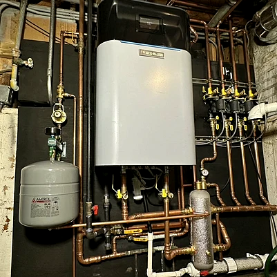 A wall-mounted boiler with copper piping, an expansion tank and zone valves, mounted on a black panel in a basement