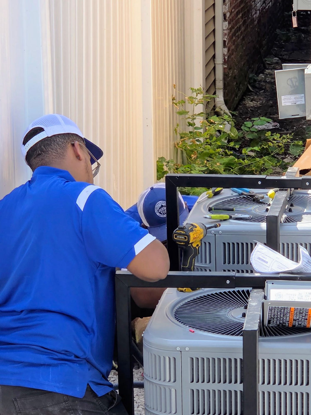 Two Mountain Top technicians in company shirts and caps installing new outdoor air conditioner condensers on a steel stand beside a building