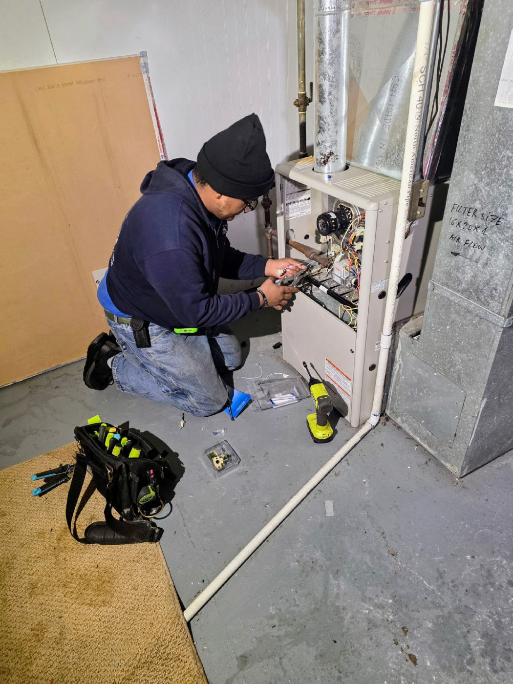 A Mountain Top technician kneeling at an open gas furnace, repairing the burner area with pliers, with his tools laid out on the floor