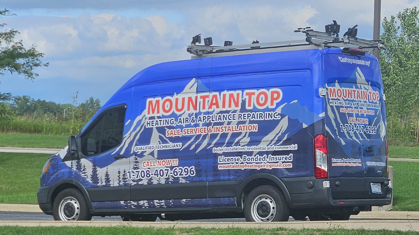 The blue Mountain Top service van, marked with the company name, phone number and licensed, bonded and insured lettering, parked in front of a building and a row of flags